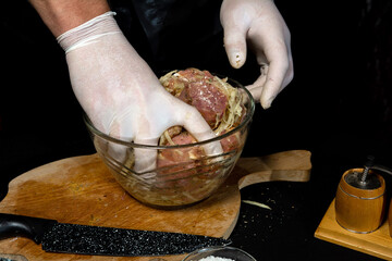 Chef in gloves kneads meat on a plate for cooking kebabs in Georgian. Pork marinade with grated potatoes and onions on a dark background.