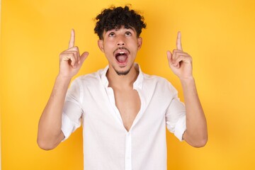 Charming carefree man with positive expression, points up with both index fingers, dressed in casual clothing, has broad intrested smile, isolated over pink background. Look there, pleaseâ€ 