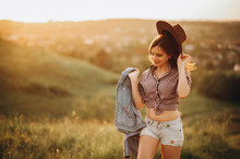Young Girl In Hat And Denim Shorts Walks Around The Field At Sunset