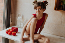 Cute Barefoot Girl In Red Bodysuit Sitting On Counter With Tomatoes And Red Pepper In Kitchen