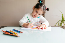 Girl In Casual Clothes Sitting On Chair At Wooden Table And Drawing Colorful Rainbow With Pencil On Paper