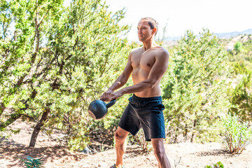 Young fit shirtless man exercising with heavy kettlebell doing swing exercise and muscles in outdoors outside park holding weight lifting