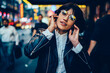 © BullRun - Brunette stylish hipster girl in optical eyewear enjoying listening electronic music in earphones standing in night downtown of New York, half length portrait of millennial tourist in headphones