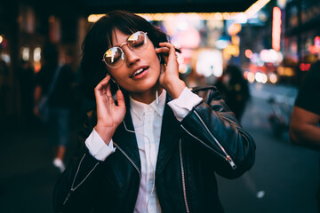 Wall Mural - Carefree millennial party girl in electronic headphones relaxing with tranquil music podcast listening in night New York, female 20s in trendy glasses and headphones enjoying media set records