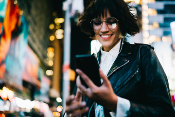 Wall Mural - Cheerful hipster girl in eyewear using mobile application for navigating in night megalopolis listening music in earphones,happy young woman reading SMS spending time in evening city