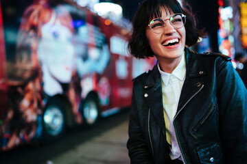 Wall Mural - Cheerful hipster girl in design spectacles laughing feeling carefree on free time walking in downtown with night lights.positive young woman in stylish wear having fun in evening megalopolis