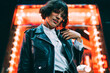 © BullRun - Half length portrait of attractive hipster girl dressed in fashion apparel listening music in earphones from cellular in downtown with night lights.Stylish female meloman standing in neon illumination