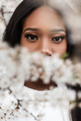 Wall Mural - Close-up portrait of a delightful young black woman with an amazing look at the camera near the flowers. Pretty girl with an expressive look looks at the camera. Beauty lifestyle