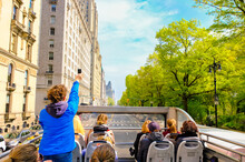 Tourists Enjoying Views Of New York City From Top Of Open Roof Of Double Decker Bus In Spring; Woman In Blue Is Taking Photos With Her Smartphone
