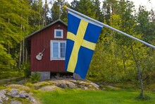 Swedish Flag Blue With Yellow Cross Waving In The Wind With A Wooden House In The Background