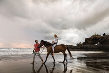 Asia, Indonesia, Bali, Young Caucasian Man With A Beautiful Young Caucasian Woman Sitting On A Horse, On A Beach At Sunset, With Typical Balinese Rock Formation Situated In The Sea