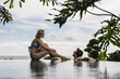 © Asia Pics - Asia, Indonesia, Bali, young beautiful Caucasian couple sitting on edge of infinity pool, with tropical plants and with the sea in the background.