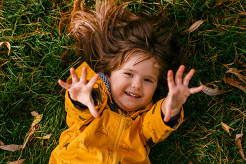 Wall Mural - Adorable little girl 3-4 years old lying on the grass on an autumn day. Cute girl-toddler resting and having fun in the Park. Close-up. The view from the top. Thick beautiful hair and yellow jacket.