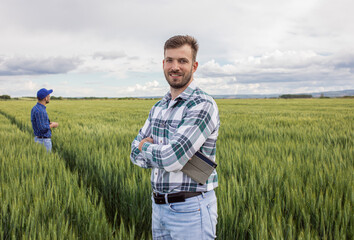 Wall Mural - Portrait of farmer standing in green wheat field with his colleague in background.