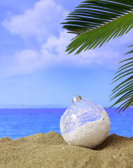 Summer xmas holidays concept. Christmas ball on sandy beach with palm tree, blue sea and sky background