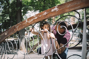 Wall Mural - Father and his daughter holding on fence in park. Cute little child and her dad standing behind decorative metal fence in city park.