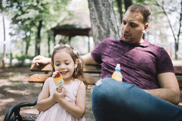 Wall Mural - Portrait of little girl holding ice cream in nature. Happy little girl holding ice cream while sitting on bench in park next to her dad.