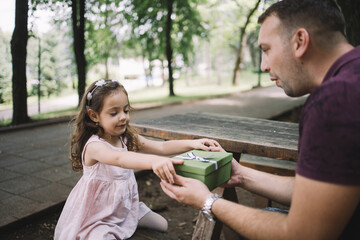 Wall Mural - Little child receiving present from father outdoor. Cute little daughter and her father resting on wooden seat in park while holding present.