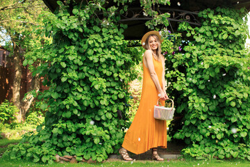 Young smiling girl in hat and orange dress in the summer green garden with basket, photography for blog and advertising