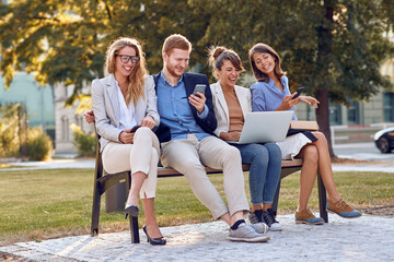 Wall Mural - young businesspeople laughing at park sitting on  a bench with their cell phones, laptop, tablet. outdoor good mood break concept