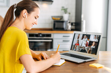 Side View Of Young Woman Watching On Laptop Display With A Group Of Multiracial People On It And Writing Notes. Video Call, Online Conference, Webinar