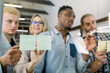 © sofiko14 - Young group of four multiethnic people standing behind glass wall in office with stickers on it and collaborating on new project. Focus on stickers and charts on glass wall, view through the glass