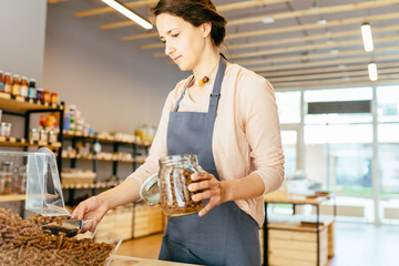 Wall Mural - Shop assistant woman owner filling reusable glass jar with dried pasta in organic grocery store. Young shopkeeper working in zero waste shop.