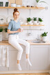© dikushin - Cheerful redhead young woman holding cup of hot coffee while standing in kitchen room with white modern interior, looking at the camera. Smiling business lady drinking tasty beverage from mug at home.