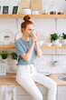 © dikushin - Pretty business lady drinking tasty beverage from mug at home office in during break. Happy redhead young woman holding cup of hot coffee while standing in kitchen room with white modern interior.