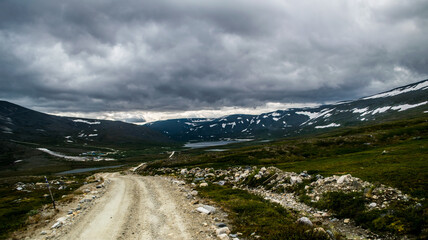gravel road to mountain ridge with small coniferous trees during stormy weather with gray clouds