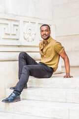 Young African American Male College Student with beard, wearing green short sleeve shirt, black pants, leather shoes, sitting on stairs of office building on campus in New York City, relaxing..