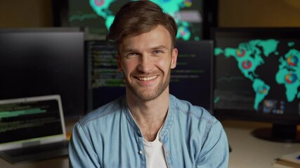 Portrait of happy handsome young man looking at camera and smiling with multiple computer monitors with programming code and global market data on screens in background in dark office