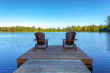 Two Muskoka chairs sitting on a wood dock facing a lake in a calm autumn season sunny day, with the chairs in the sun shadow.