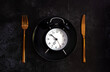 © Jane Vershinin - Alarm clock on a black plate with copper knife and fork. Intermittent fasting concept