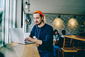 Fototapete - Caucasian male resting in coffee shop listening favourite music in headphones and chatting in social networks, skilled freelancer enjoying remote job typing publication for web blog on laptop