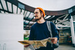Happy Caucasian male tourist in stylish glasses smiling during travel sightseeing for exploring city streets, good looking backpacker holding paper map for search location direction and walking