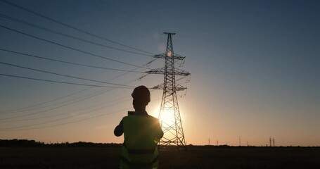 Wall Mural - worker in uniform inspects power lines using a tablet on sunset background.