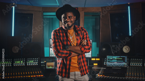 Portrait of Young Black Artist, Musician, Audio Engineer, Producer Wearing Stylish Hat and Standing in Music Record Studio. In the Background Control Desk, Mixing Equipment, Soundproof Room.