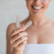 Close-up of orthodontic silicone transparent teeth aligner in female hands. Blurred unrecognizable woman in a white towel holds a removable night retainer. Brace for teeth whitening.