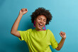 © wayhome.studio  - I am winner! Ecstatic overjoyed African American woman dances carefree, celebrates victory and success, dressed in green casual t shirt, feels lively and energetic, isolated on blue background