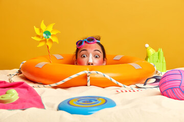 Photo of surprised beautiful woman with eastern appearance, relaxes on beach looks stunned at camera, head over sand inflated lifebuoy around spends summer vacation at seaside sunbathes with sunscreen