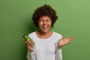 Wall Mural - Happy healthy woman laughs from joy, holds glass bottle of green vegetable smoothie, cares about figure, has proper nutrition, dressed casually, isolated on bright vivid background. People, lifestyle