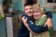 Happy, smiling grandson hugs his grandmother in the kitchen at home. Happy family relationships, love, care. Poor, real people.