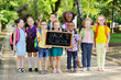 © Evgeniy Kalinovskiy - a group of multi-racial school children in colorful clothes, carrying school bags and backpacks hold a sign that reads 'Back to school' against the background of greenery and a Park.