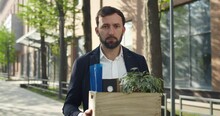 Portrait of young man in suit holding box with his stuff while standing at street. Male office worker in 30s lost his job and looking disappointed. Concept of unemployment and crisis