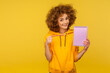 © khosrork - Portrait of cheerful curly-haired woman in urban style hoodie showing pencil and paper notebook to write idea, looking at camera with toothy smile. indoor studio shot isolated on yellow background