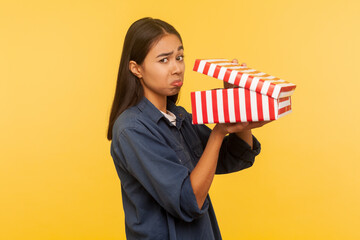 Portrait of upset girl in denim shirt opening gift and looking at camera with unhappy disappointed expression, frustrated emotions, confused by wrong delivery. indoor studio shot, yellow background