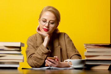 Wall Mural - blonde woman sitting at table with books and using mobile phone on yellow background