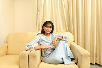 Wall Mural - young Asian Malay Muslim woman wearing baju kurung dress at home sit rest on sofa with feets up