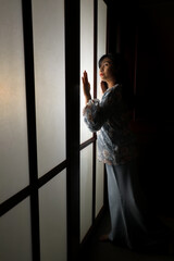 Wall Mural - young Asian Malay woman wearing baju kurung dress at home in front window mood light on face dark background peaceful content calm tranquil break trough wonder